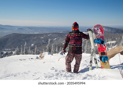 A Man In Snowboarding Gear On The Top Of A Mountain. Young Man Standing With A Snowboard On The Top Of The Mountains.