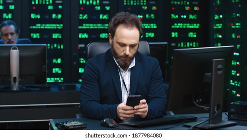 Man in solid suit sitting in office at work on background of working servers holding smartphone flipping through the news on social networks.