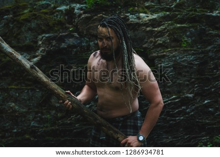 A man with soot on his face and in a Celtic skirt carries a charred log in the woods near the rock
