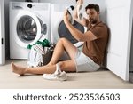 man sorting clothes near washing machine. man sits on the floor of a house near a washing machine with dirty clothes.man holding a dirty sock sitting by the washing machine, dirty shoes, dry cleaner.