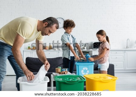 Man sorting garbage near blurred kids and trash cans with recycle sign at home Foto stock © 