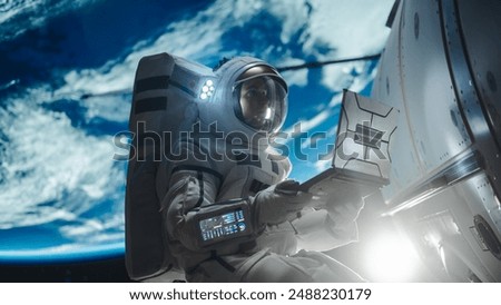 Man in a Space Suit Casually Working on a Laptop Computer in Outer Space While Servicing a Satellite. Astronaut Writing to Mission Control, Family or Work Colleagues, Reading Maintenance Protocol