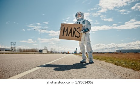 Man in Spacesuit is Standing at the Edge of a Road and Holding a Sign with Mars Written on it. Astronaut Looking to Hitchhike a Car. Spaceman in Futuristic Suit with Technological Panel on His Hand.