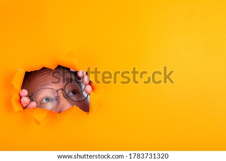 The man with spectacle peeks through a cut hole in the orange paper background.