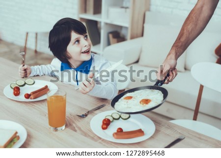 Man Spends Time with His Son. Father of Boy is Engaged in Raising Child. Father is Feeding a Eldest Son a Breakfast. Son is Sitting on Chair at Table and Smiling. People Located on Kitchen.
