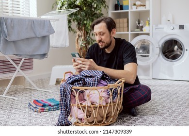 A Man Spends Time In Laundry Room While Doing Household Chores, Boy Sorting Clothes For Washing, Holding Phone In Hands, Shopping Online, Ordering Powder And Liquid For The Washing Machine.