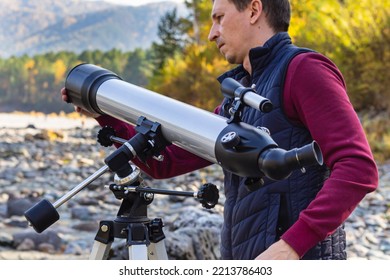 Man In Sportswear Sets Up Gray Telescope On Tripod On River Bank,scientific Tool For Space Observation. Amateur Telescope Mounted On Tri-pod In Mountain Valley, Look At Sky With Special Lenses.