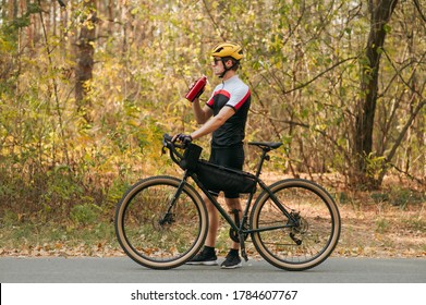 Man in sportswear standing in autumn forest with bicycle and bottle of water in his hands and looking away. Athlete weightlifter is resting in training, drinking water from a bottle.