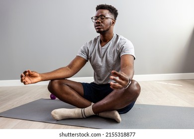 Man in sporty outfit doing yoga and meditating on an exercise mat. Sporty mindful man with tattoo meditating alone at home, peaceful calm hipster fit guy practicing yoga in lotus pose 