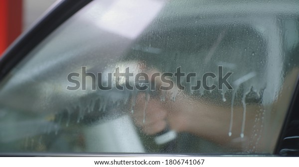 Man spraying water on\
glass then removing foam with spatula while washing vehicle window\
in garage