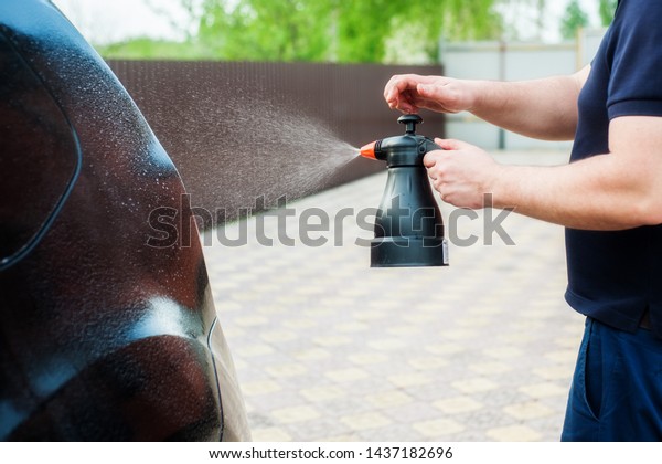 A man sprays car shampoo on a black car from\
a plastic bottle. Car wash\
concept