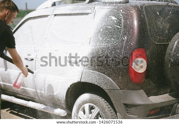 A man sprays a cleaning agent with a high pressure\
washer on a car. The car washes with a means of washing. The\
machine is in foam