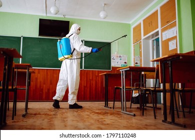 Man Sprays Disinfection Liquid Over The Desks At The Classroom Before The School Season. Sanitary Worker Wearing Protective Suit Cleans The Auditorium. Students And Pupils Health Care Concept.