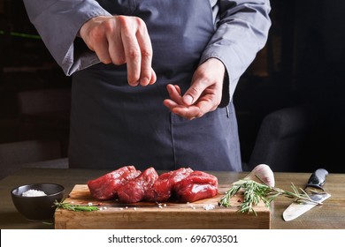 Man sprinkles filet mignon steaks with pepper salt. Chef working at open restaurant kitchen. Fresh meat, garlic and rosemary on wooden board. Modern restaurant cuisine backgroung - Shutterstock ID 696703501