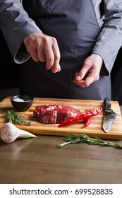 Man sprinkles rib eye steak with pepper salt. Chef working at open restaurant kitchen. Fresh meat, garlic, rosemary and red chilli on wooden board. Modern restaurant cuisine backgroung, copy space - Shutterstock ID 699528835