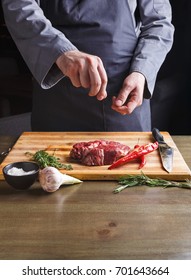 Man sprinkles rib eye steak with pepper salt. Chef working at open restaurant kitchen. Fresh meat, garlic, rosemary and red chilli on wooden board. Modern restaurant cuisine backgroung, copy space - Shutterstock ID 701643664