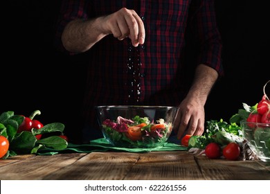 Man Sprinkles Salt Salad Of Fresh Vegetables On A Wooden Table. Preparing Healthy Food