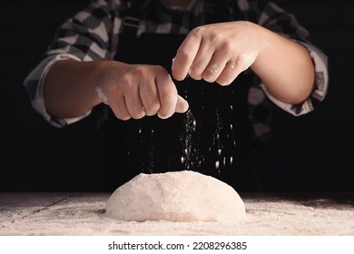 Man sprinkling flour over dough at wooden table on dark background, closeup - Shutterstock ID 2208296385