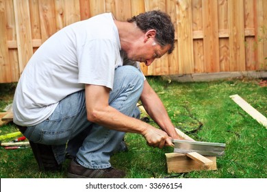 Man Squat Down Sawing A Piece Of Wood In Front Of A Wooden Fence In The Garden