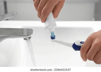 Man squeezing toothpaste from tube onto electric toothbrush above sink, closeup - Powered by Shutterstock