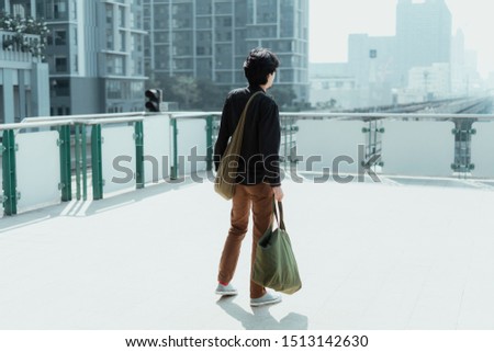 A man stading on skytrain platform carrying cloth bag.