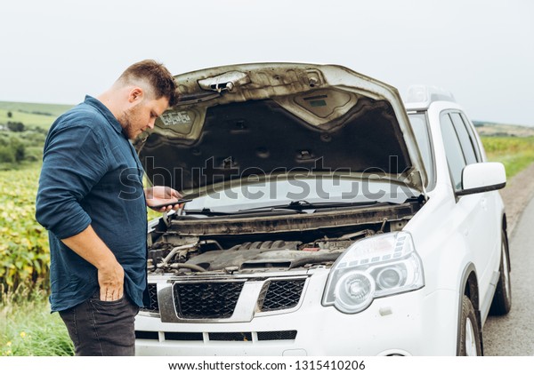 man stand in front of broken car with phone.\
broken in the middle of\
nowhere