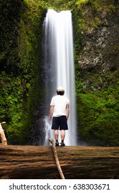 A man stand upon a fallen tree, observing the flowing beauty of a majestic waterfall and feeling one with nature.