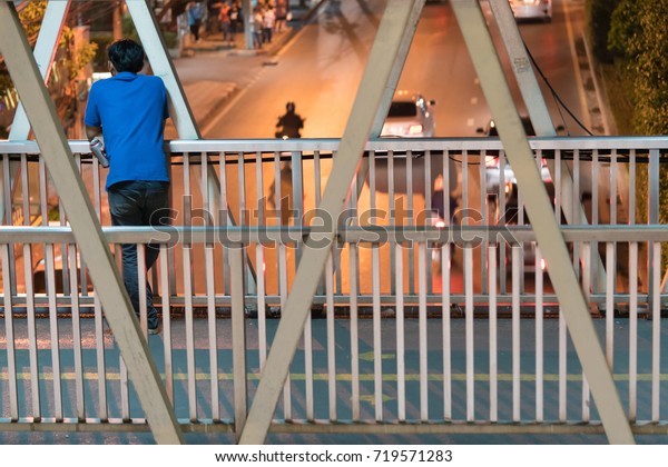 Man standing alone on the\
bridge. Asian man standing behind and holding a beer can on the\
footbridge and looking at the car run on the street, in the city\
night time.