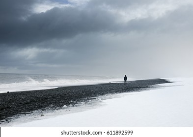 Man Standing Alone On The Dark And Snow Beach Under Cloudy Sky And Wild Sea