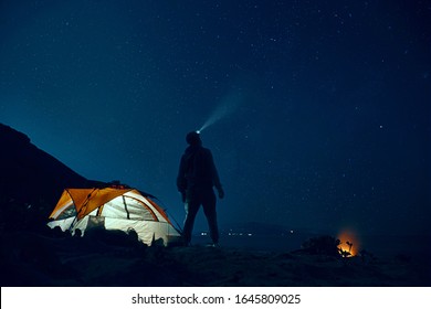 Man Standing Beside Camping Tent Wearing Headlamp During Nighttime.