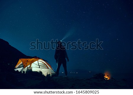 Man standing beside camping tent wearing headlamp during nighttime.