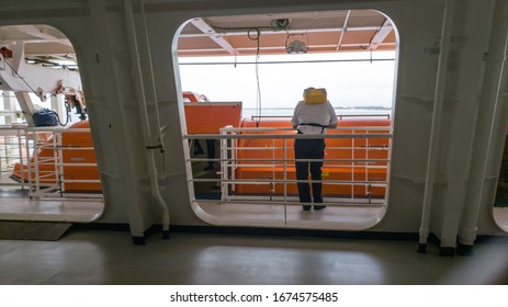 Man Standing By The Life Boat On A Cruise Ship Open Deck During COVID-19 Lockdown Wearing Yellow Life Jacket On A Safety Drill