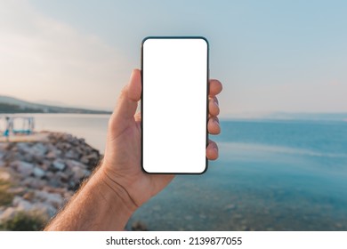 Man standing by the sea and holding mobile smart phone with blank mockup screen, closeup of hand and device, selective focus - Powered by Shutterstock