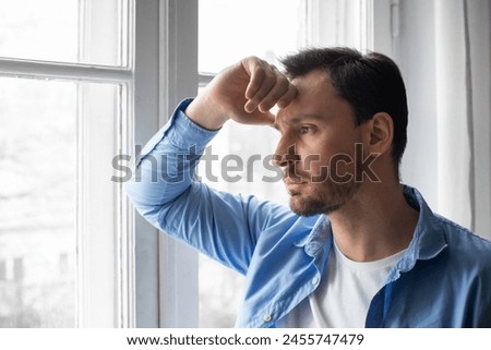 A man is standing by a window, looking outside. His hand is resting on his head as he appears lost in thought, perhaps contemplating or reflecting