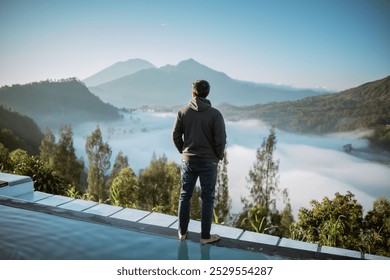 A man is standing confidently on a ledge while admiring the breathtaking view of a beautiful lake surrounded by majestic mountains - Powered by Shutterstock