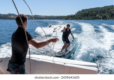 A man standing at the edge of a moving boat, receiving the rope from a wakeboarder surf behind in the lake, enjoying the summer day on the water. - Powered by Shutterstock