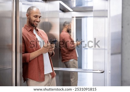 Man standing in elevator using smartphone, leaning against mirror wall, smiling effortlessly, dressed in casual attire. Scene depicts connectivity, communication, modern technology, daily routine.