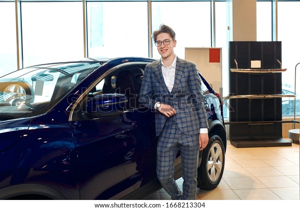 Man standing in front of a car in a showroom and\
posing to the camera, elegamt dealer looking at camera. close up\
photo. guy has bought a new car for his wife, girlfriend, positive\
feeling and emotion