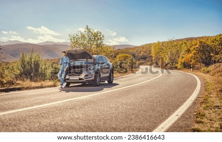 Man standing in front of an open hood of a car on an open road and typing on a smartphone, car breakdown concept