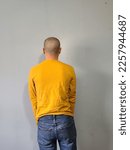 A man standing in front of a wall while wearing a long sleeved yellow shirt.