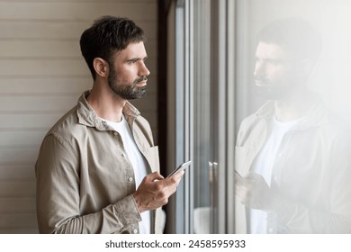 A man is standing in front of a window, looking at his cell phone. He appears focused on the screen, with natural light coming through the window.