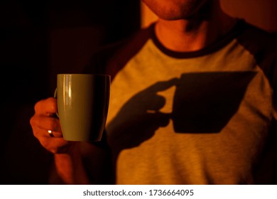 A man standing in front of a window in sunset lighting is drinking coffee in his kitchen