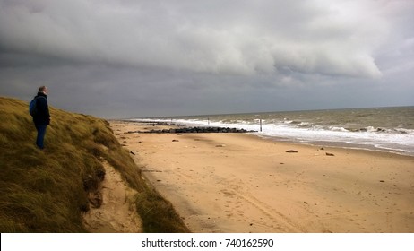 A man standing grassy sand dunes gazing out to sea on a cold windy stormy Winter day on a sandy beach in Norfolk East Anglia England with rough waves and grey cloudy sky - Powered by Shutterstock