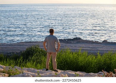 A man standing with his back on a cliff on the beach is watching the sea. man's hands in pockets and he is thoughtful. - Shutterstock ID 2272410625
