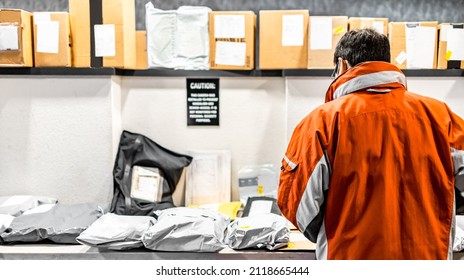 Man Standing At Mailroom Inside Apartment Looking For Packaging. Messenger Deliver Parcel Or Receiver Concept. Guy In Red Uniform Jacket With Pile Of Delivering Bags And Boxes At Distributor Storage.