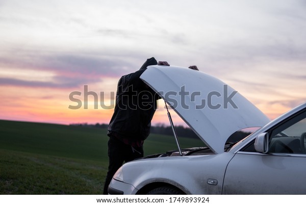 Man\
standing near car with open hood on sunset. Car breakdown. Confused\
man stands with open hood with selective soft\
focus.