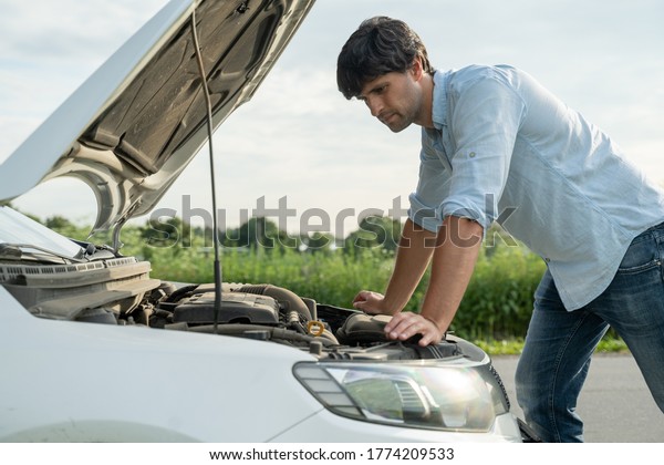 Man standing next to a broke down car,\
looking down at engine in\
frustration