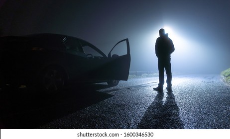 A Man Standing Next To A Car, With Door Open, Parked On The Side Of The Road, Underneath A Street Light, On A Spooky, Scary, Rural, Country Road. On A Foggy Winters Night