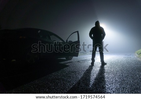 A man standing next to a car, with door open, parked on the side of the road, underneath a street light. On a foggy winters night