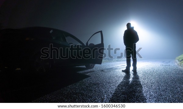 A man standing\
next to a car, with door open, parked on the side of the road,\
underneath a street light, on a spooky, scary, rural, country road.\
On a foggy winters night